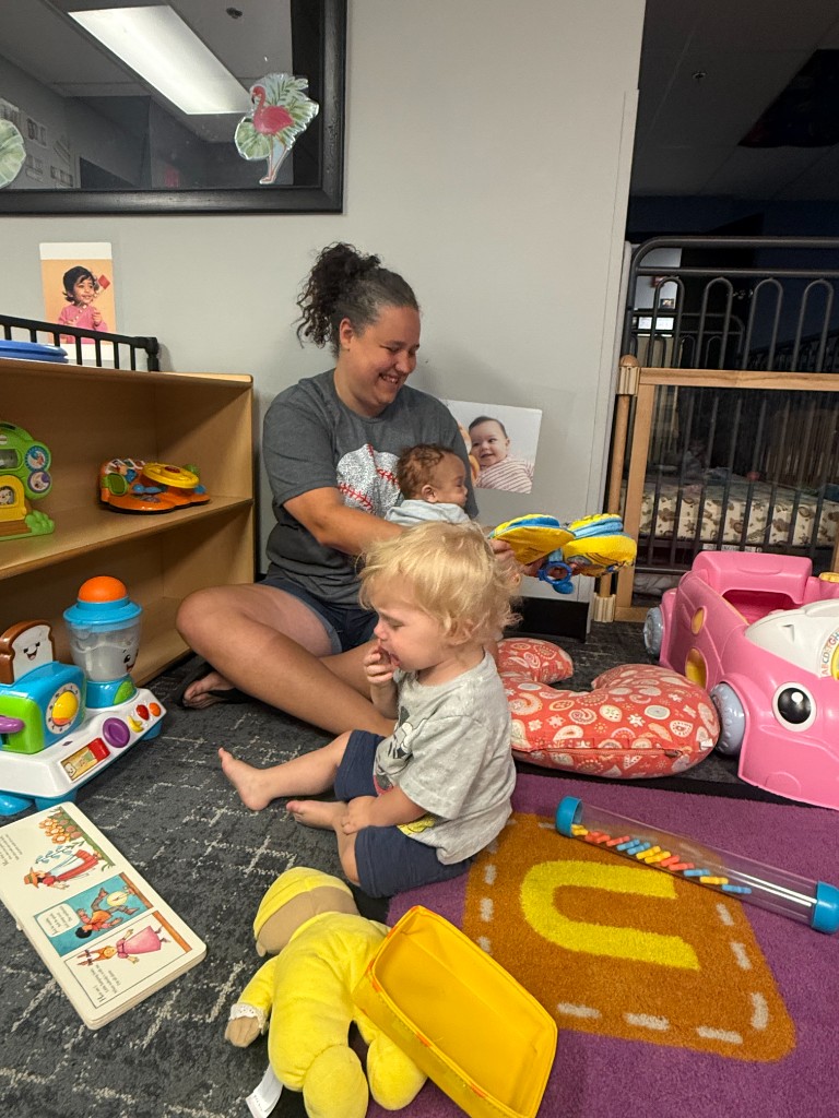 A caregiver with an infant and toddler surrounded by toys in the classroom